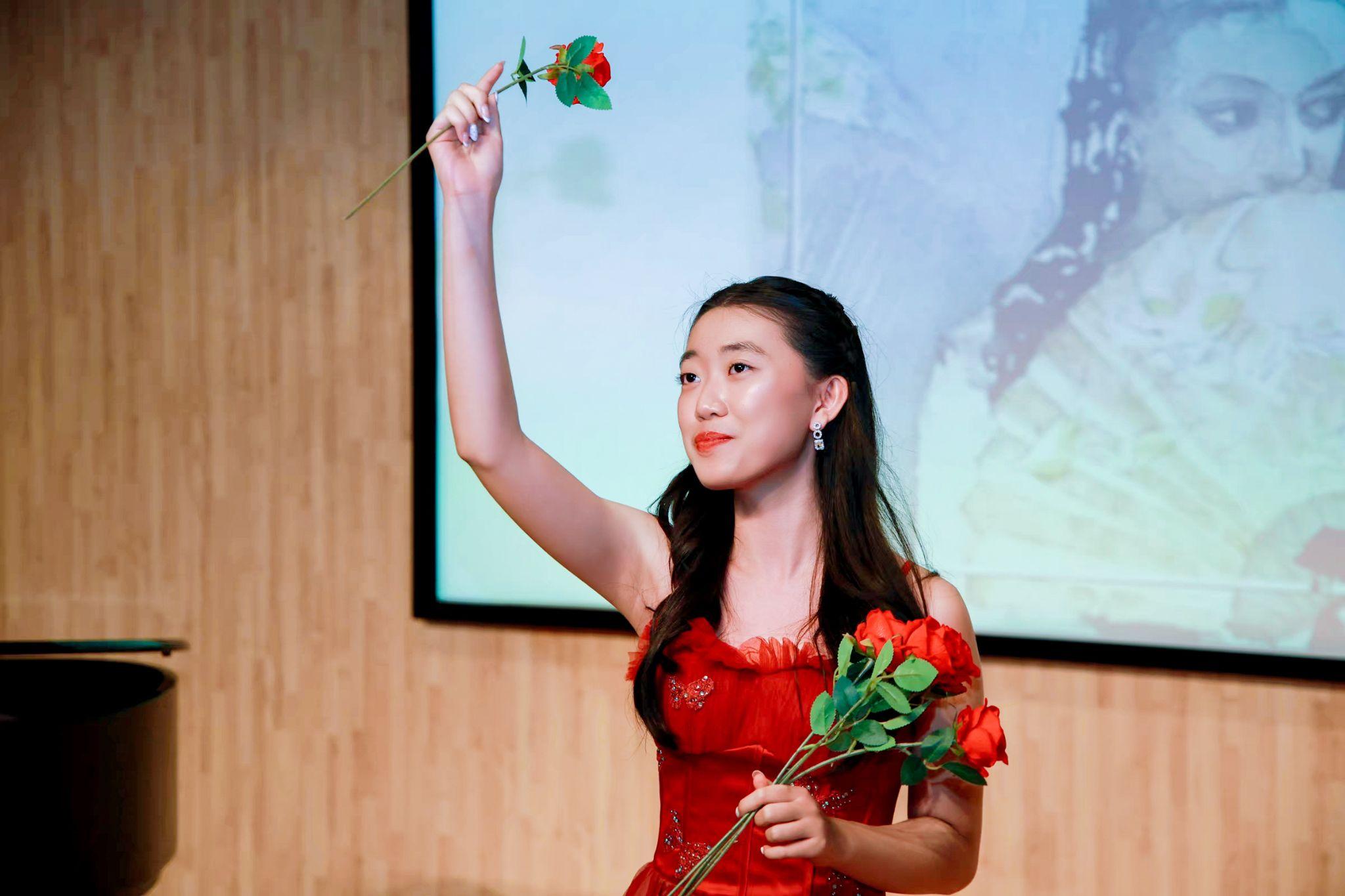 Seven Jing performing on stage in a red gown, holding a single red rose aloft and a bouquet of roses in the other hand.