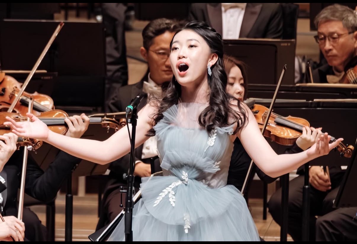 Seven Jing singing on stage in a pale blue gown, arms outstretched, in front of an orchestra's violin section.